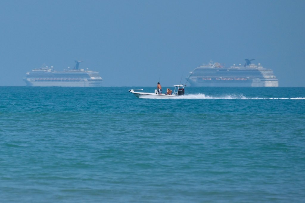 A small fishing boat on the ocean with two carnival cruise ships on the horizon