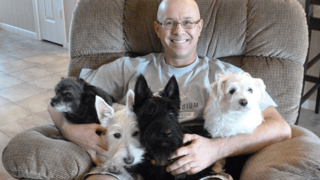 Picture of Eric Larkin on a recliner with 4 dogs on his lap. two of the dogs are scotties and two are chihuahua mixes.