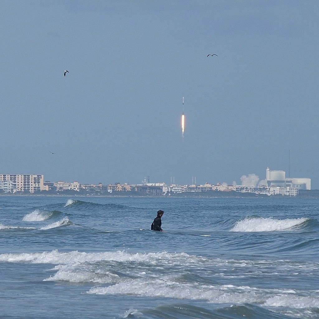 Beach photo of a spaceX launch wth a surfer in the water