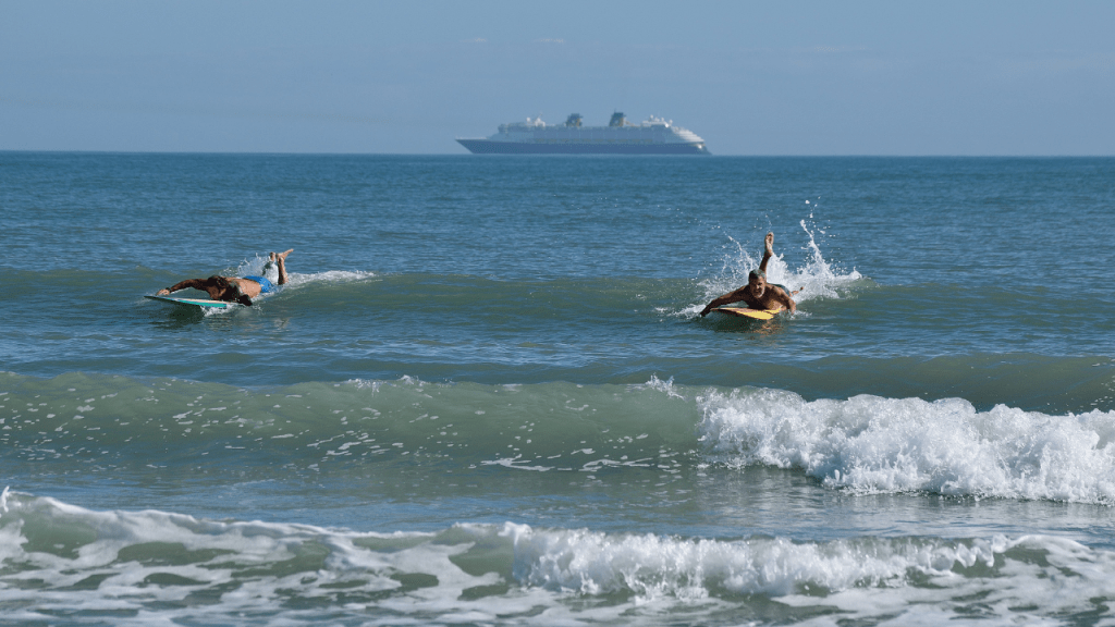 two surfers paddling to catch a wave with the disney cruise ship in the background